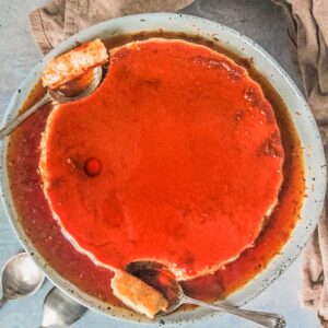 Spoonfuls of flan rest beside a large flan sitting on a rimmed blue ceramic plate above a gray surface.