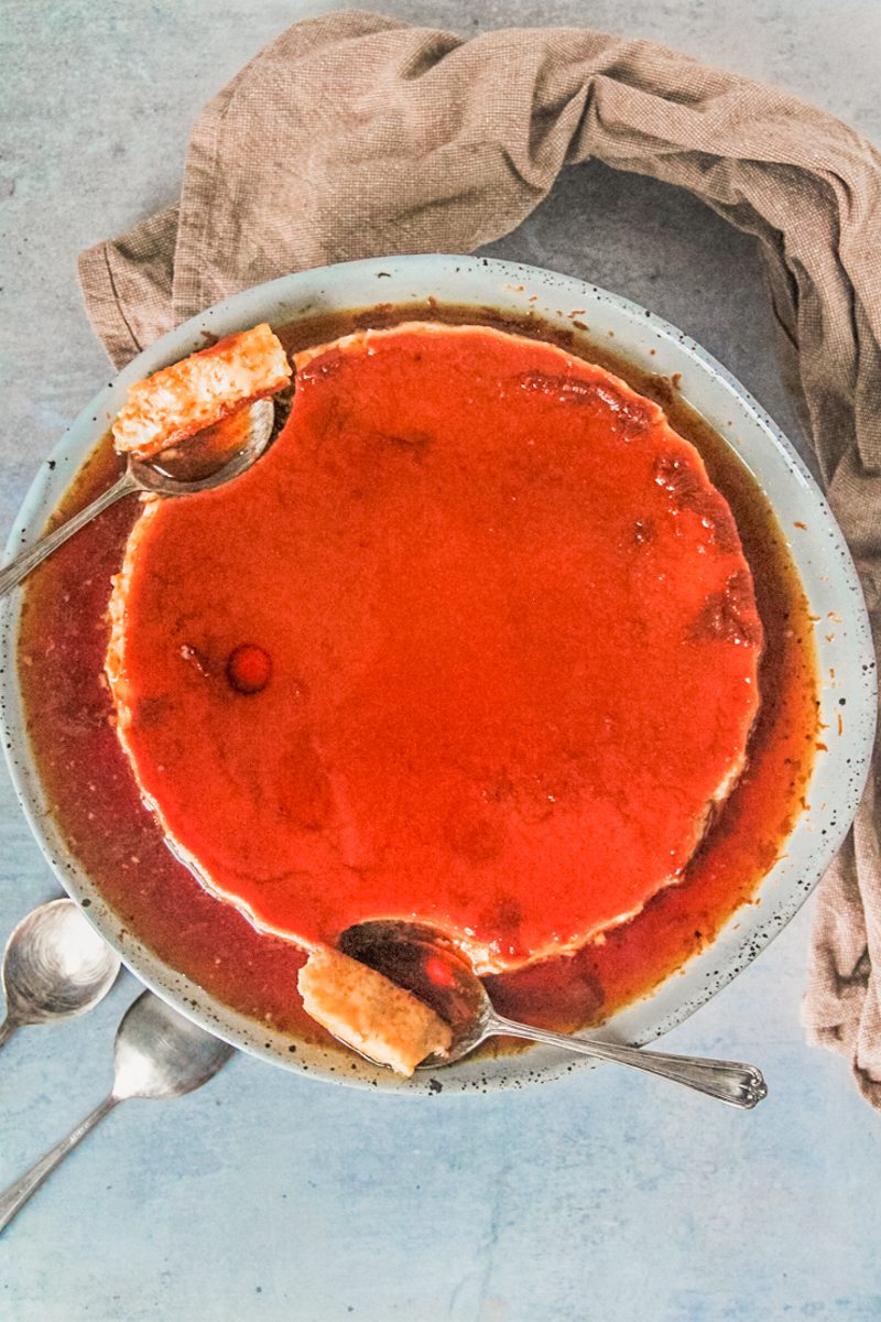 Spoonfuls of flan rest beside a large flan sitting on a rimmed blue ceramic plate above a gray surface.