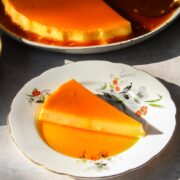 A slice of Mexican Flan sits on a flowery ceramic plate with caramel sauce surrounding it with the remaining flan sitting behind on a gray surface.