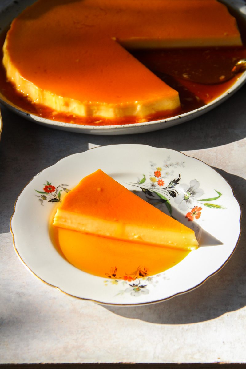 A slice of Mexican Flan sits on a flowery ceramic plate with caramel sauce surrounding it with the remaining flan sitting behind on a gray surface.