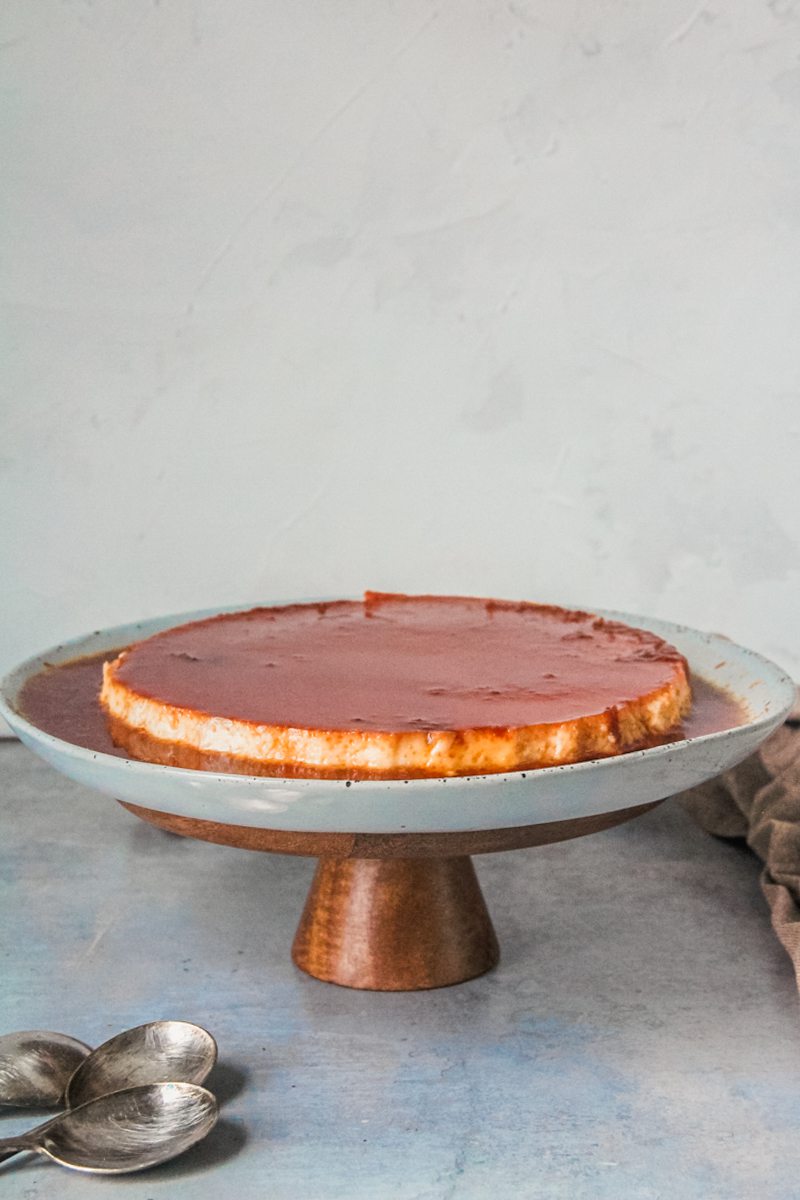 Flan sits in a rimmed ceramic plate on a cake stand on a gray surface.