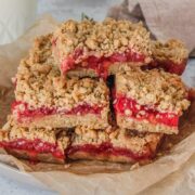 Oatmeal strawberry bars sit stacked on a rimmed plate with pieces of parchment paper on a light gray background.