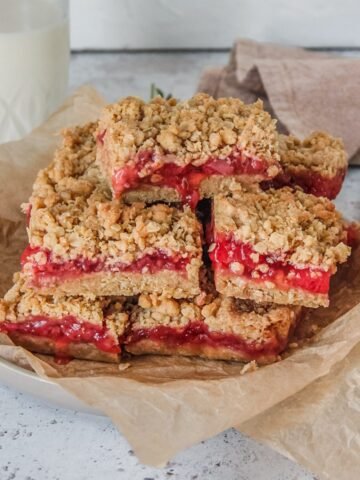 Oatmeal strawberry bars sit stacked on a rimmed plate with pieces of parchment paper on a light gray background.