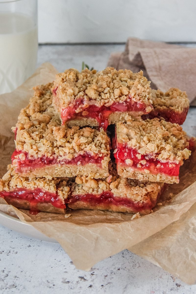 Oatmeal strawberry bars sit stacked on a rimmed plate with pieces of parchment paper on a light gray background.