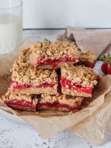 French Strawberry Granola Bars sit stacked on a plate with the juicy strawberry filling running down the oatmeal crumb base, sitting on a light grey background.