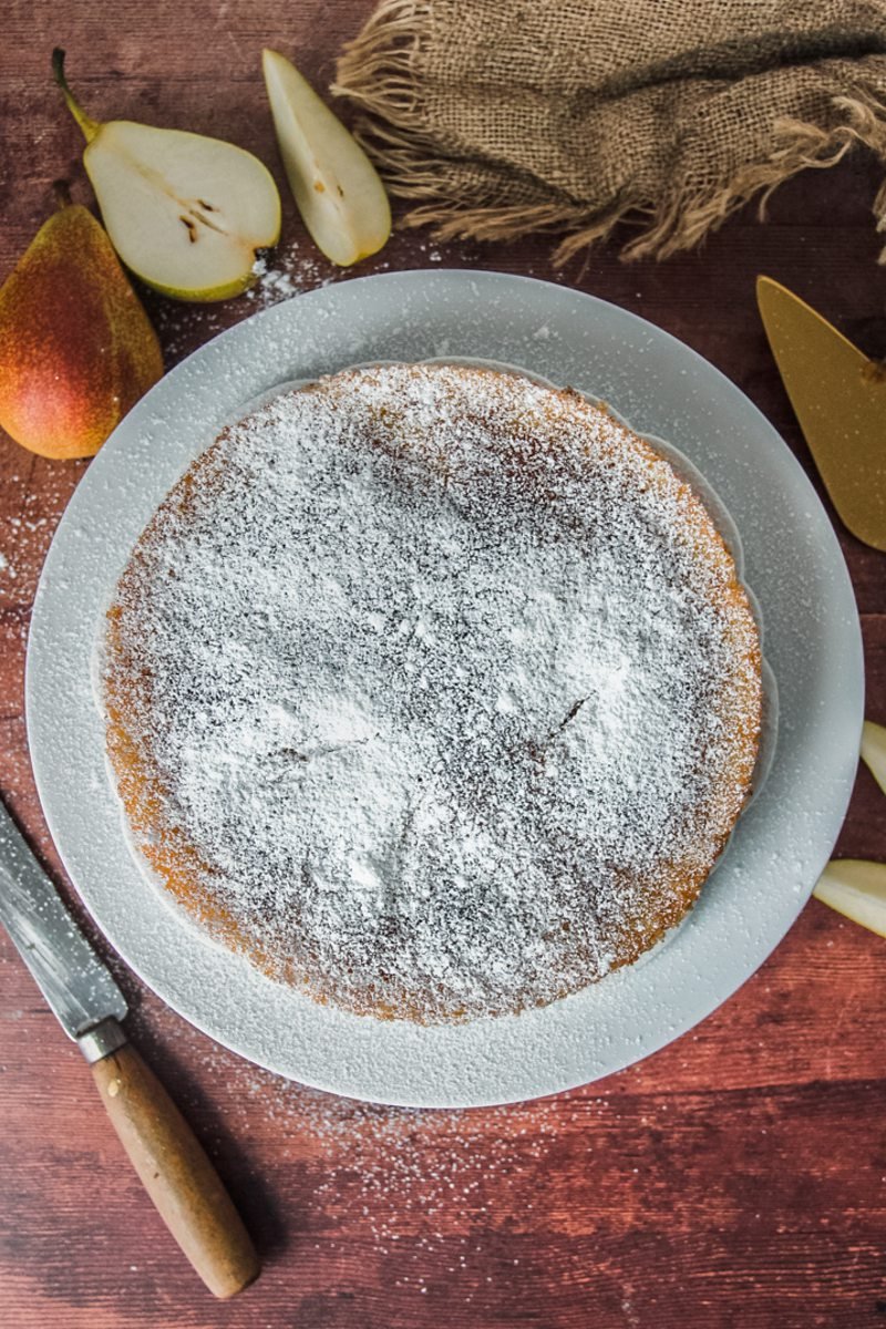 A French pear cake topped with a dusting of powdered sugar sits on white plates on a wooden surface with sliced pears on the side.