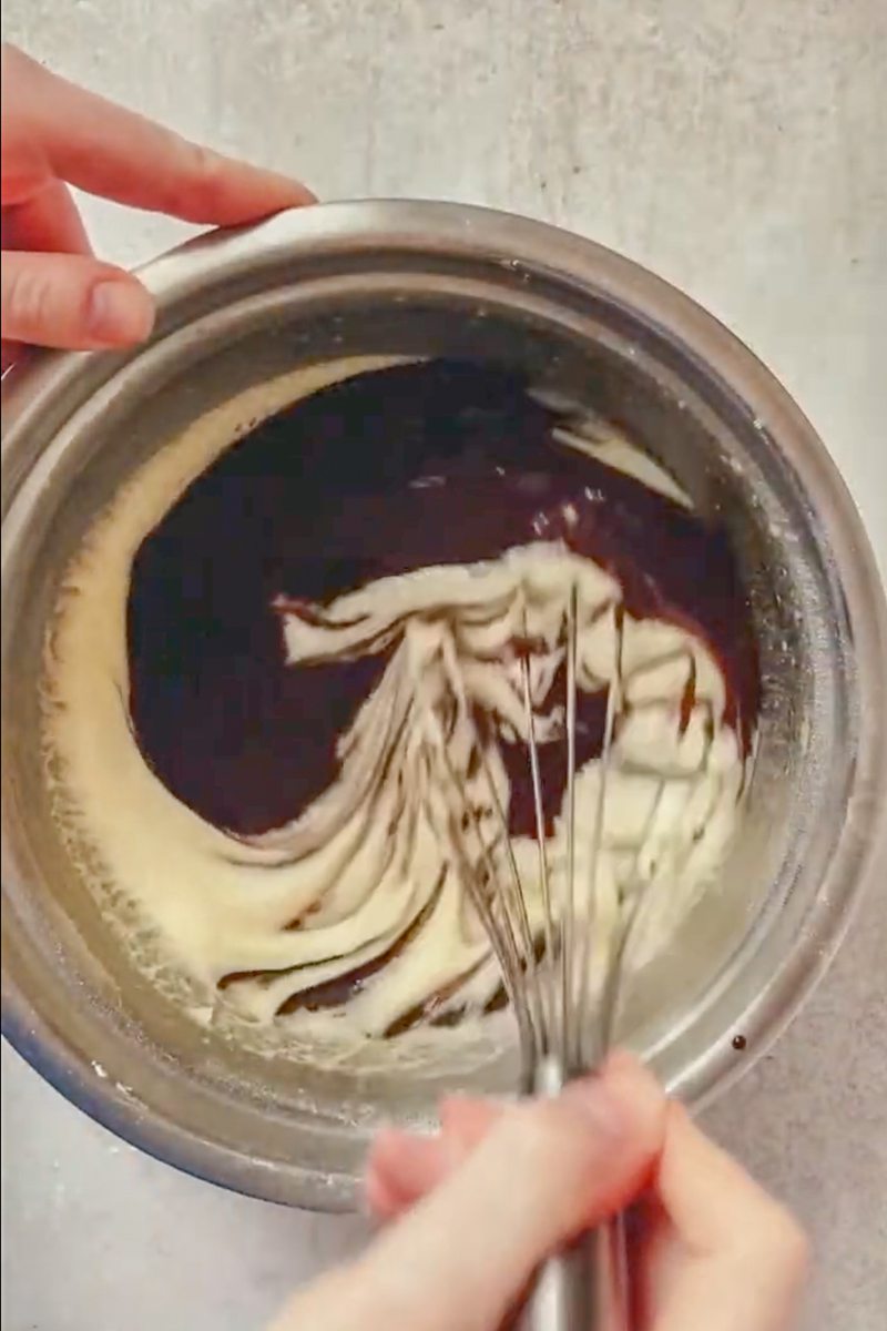 Melted butter and chocolate are whisked into a whipped egg mixture in a stainless steel bowl on a gray surface.