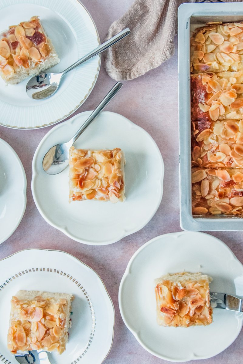 Slices of German Butter Cake sit on individual white ceramic plates beside a tray of remaining slices on a light pink surface.
