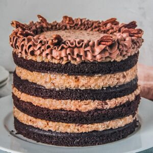 A four layered German chocolate cake with a pecan coconut filling and border of chocolate buttercream sits on a white ceramic plate on a light gray surface.