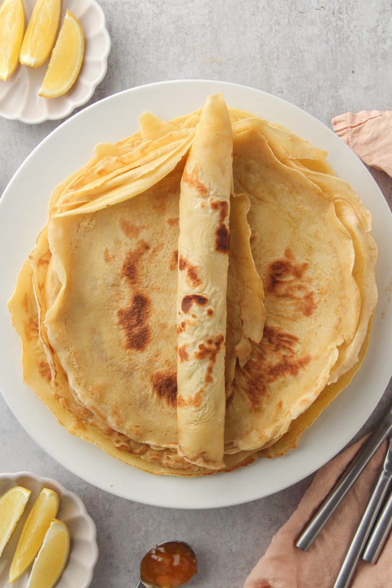 A rolled up German pancake sits on top of a stack of other pancakes on a white ceramic plate on a gray surface with lemon wedges beside.