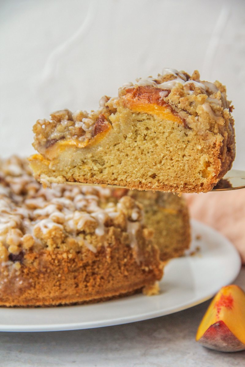 A slice of Peach Crumb Cake sits on a cake server above the remaining cake sitting on a round white ceramic plate on a gray surface.