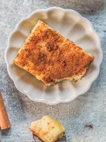 A slice of Greek Galatopita sits on an individual fluted ceramic plate on a gray surface with a spoonful of pudding beside.