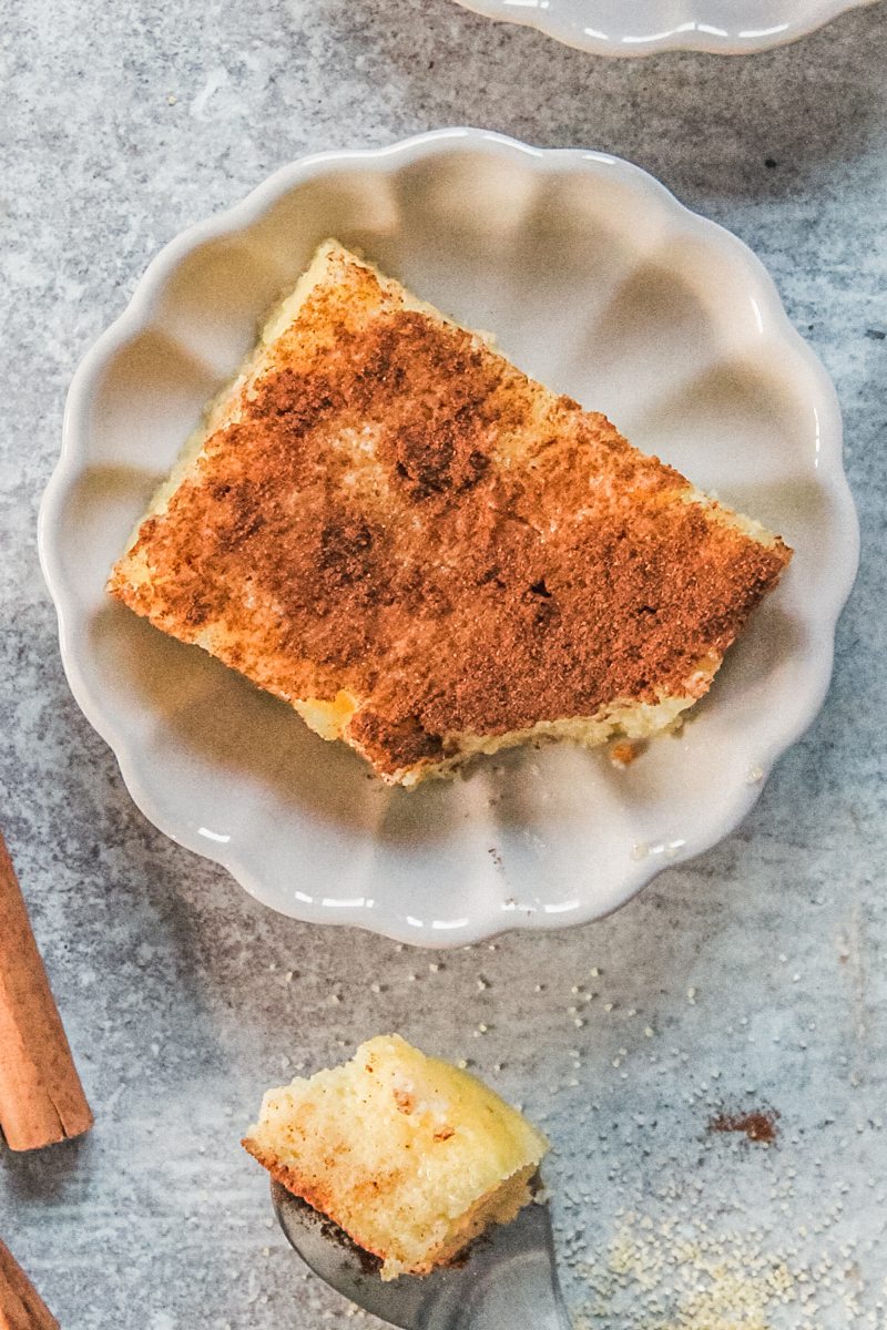 A slice of Greek Galatopita sits on an individual fluted ceramic plate on a gray surface with a spoonful of pudding beside.