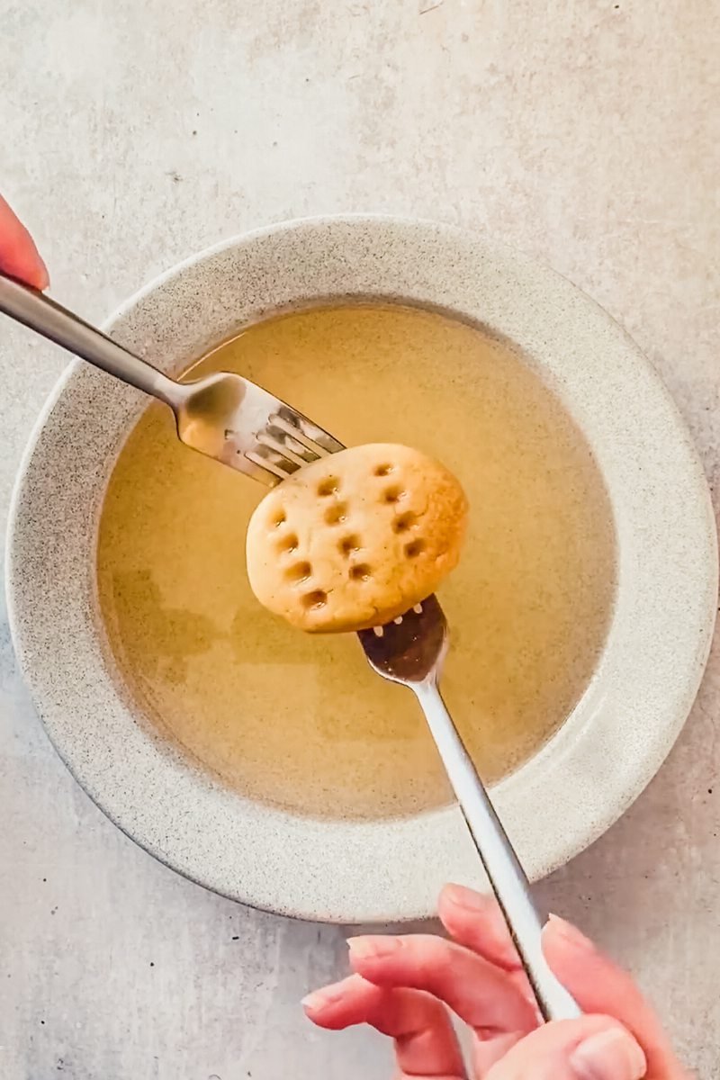 Greek holiday cookies are drained of syrup on two forks above a gray ceramic soup bowl on a gray surface.