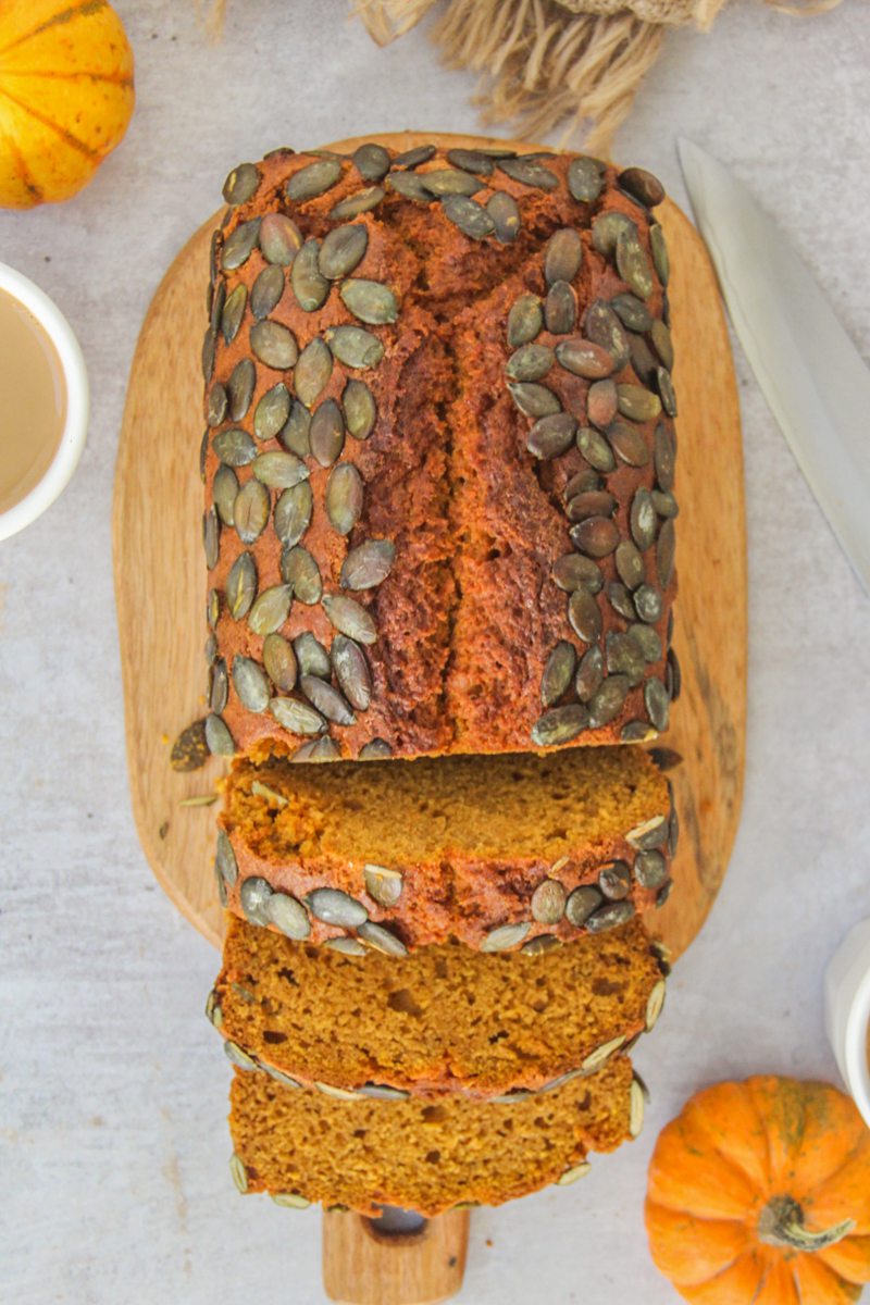 A loaf of pumpkin bread with a few slices resting; sits on a wooden board on a gray surface with small pumpkins beside.