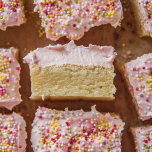 Sugar cookie bars sit on a parchment paper with a raspberry buttercream frosting topped with hundreds and thousands sprinkles and one on it's side with the soft sugar cookie bar and fluffy frosting visible.