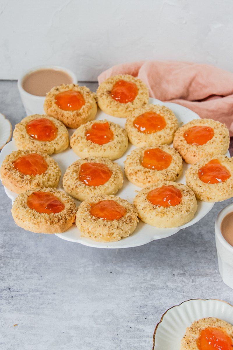 Hungarian Thumbprint cookies sit on a white ceramic plate on a light gray surface with two cups of tea beside.