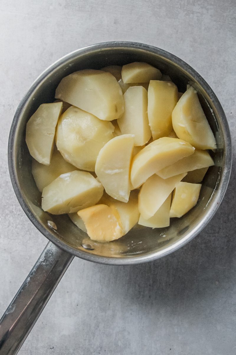 Roughly chopped cooked potatoes sit in a stainless steel saucepan with butter and milk on a gray surface.