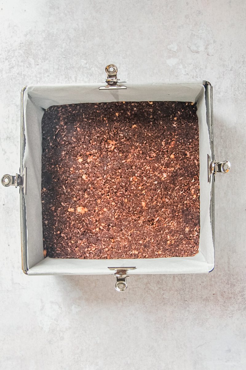 Nanaimo bar crushed biscuit and cocoa base sits pressed into a lined square metal baking tin on a gray surface.