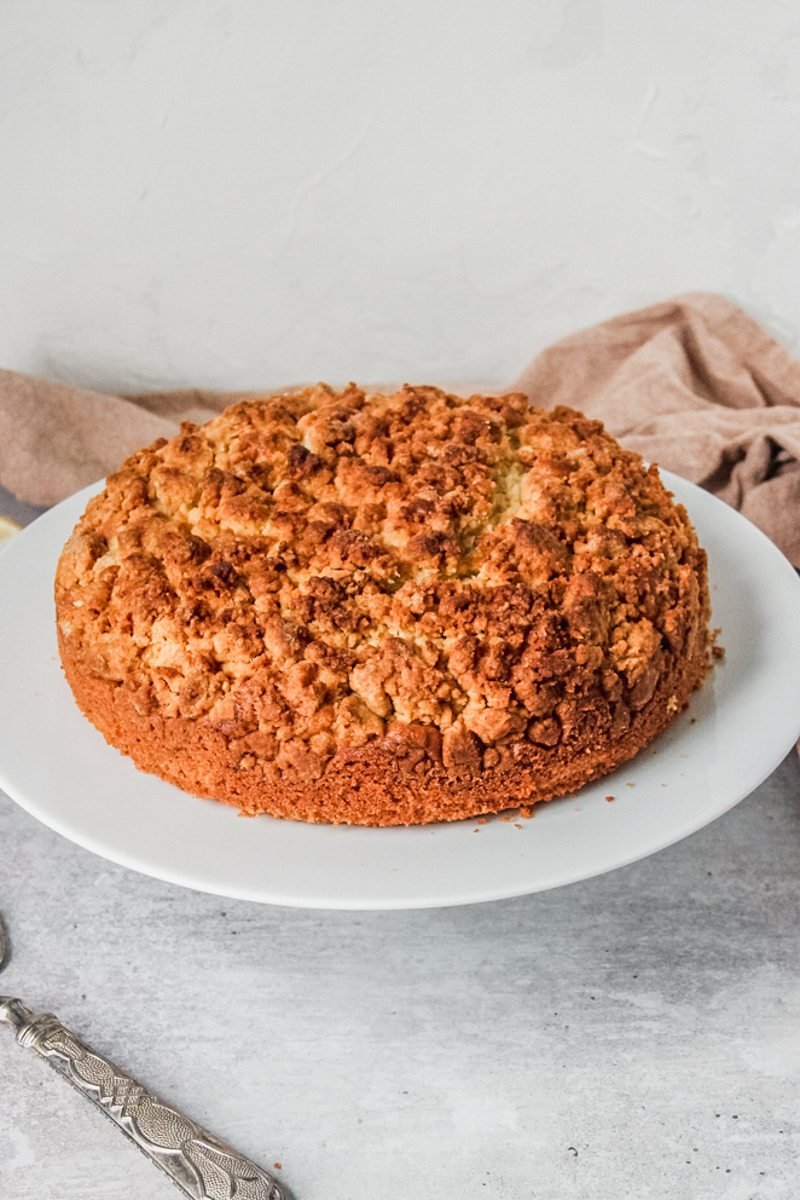 Olive oil crumble cake sits on a white ceramic plate on a cake stand on a gray surface.