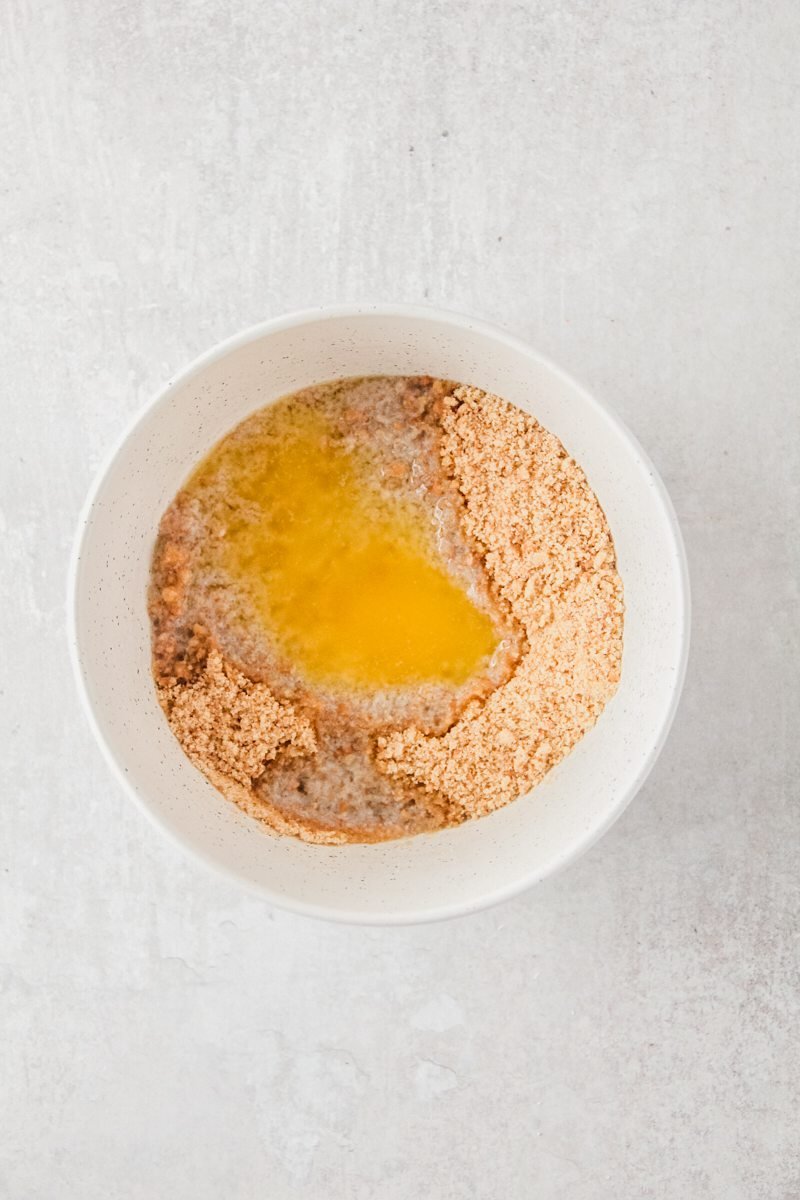 Melted butter soaks into crushed biscuits in a white ceramic bowl on a gray surface.