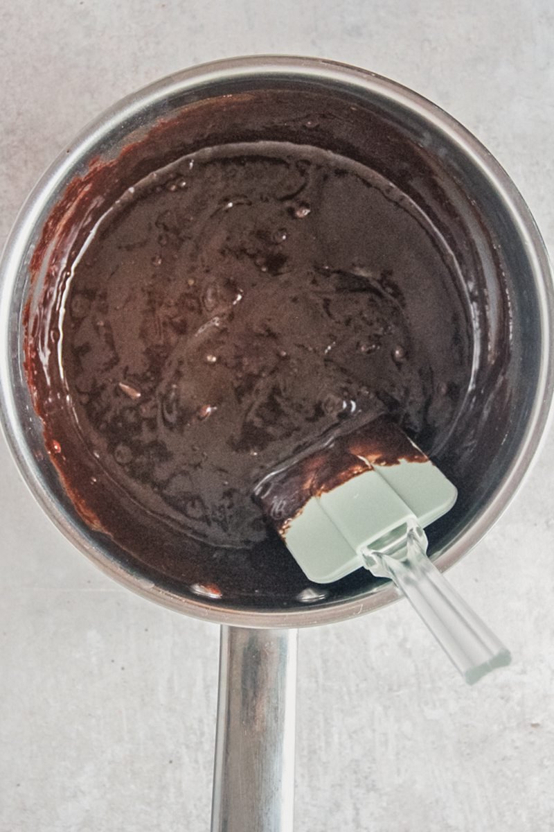 A chocolate mixture sits in a stainless steel saucepan on a gray surface with a spatula resting in the mixture.