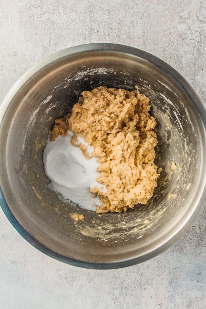 Sugar sits beside a rough dough in a stainless steel bowl on a gray surface.