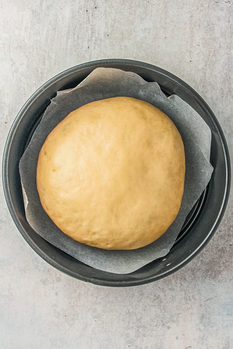 A raised smooth dough sits in a lined cake tin on a gray surface.