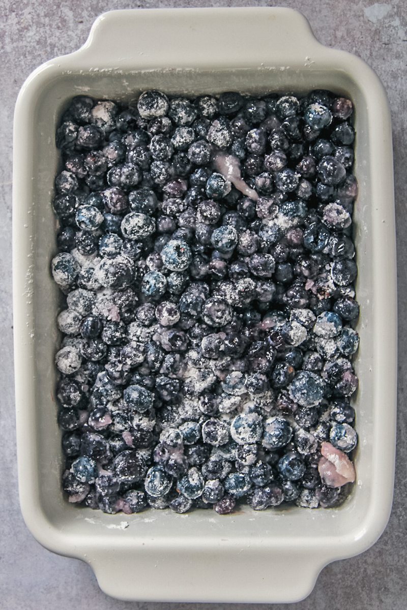 Blueberries coated in sugar and cornstarch sit in a rectangle white ceramic baking dish on a gray surface.