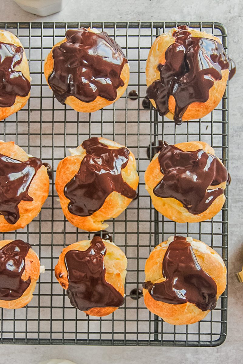 A cooling rack has cream puffs atop with a glossy chocolate sauce on top above a gray surface.