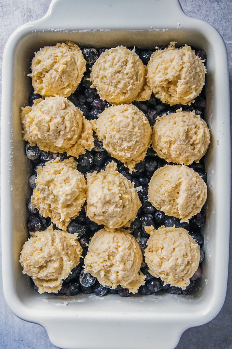 Scoops of cobbler dough sit on top of blueberries in a rectangle white ceramic baking dish on a gray surface.