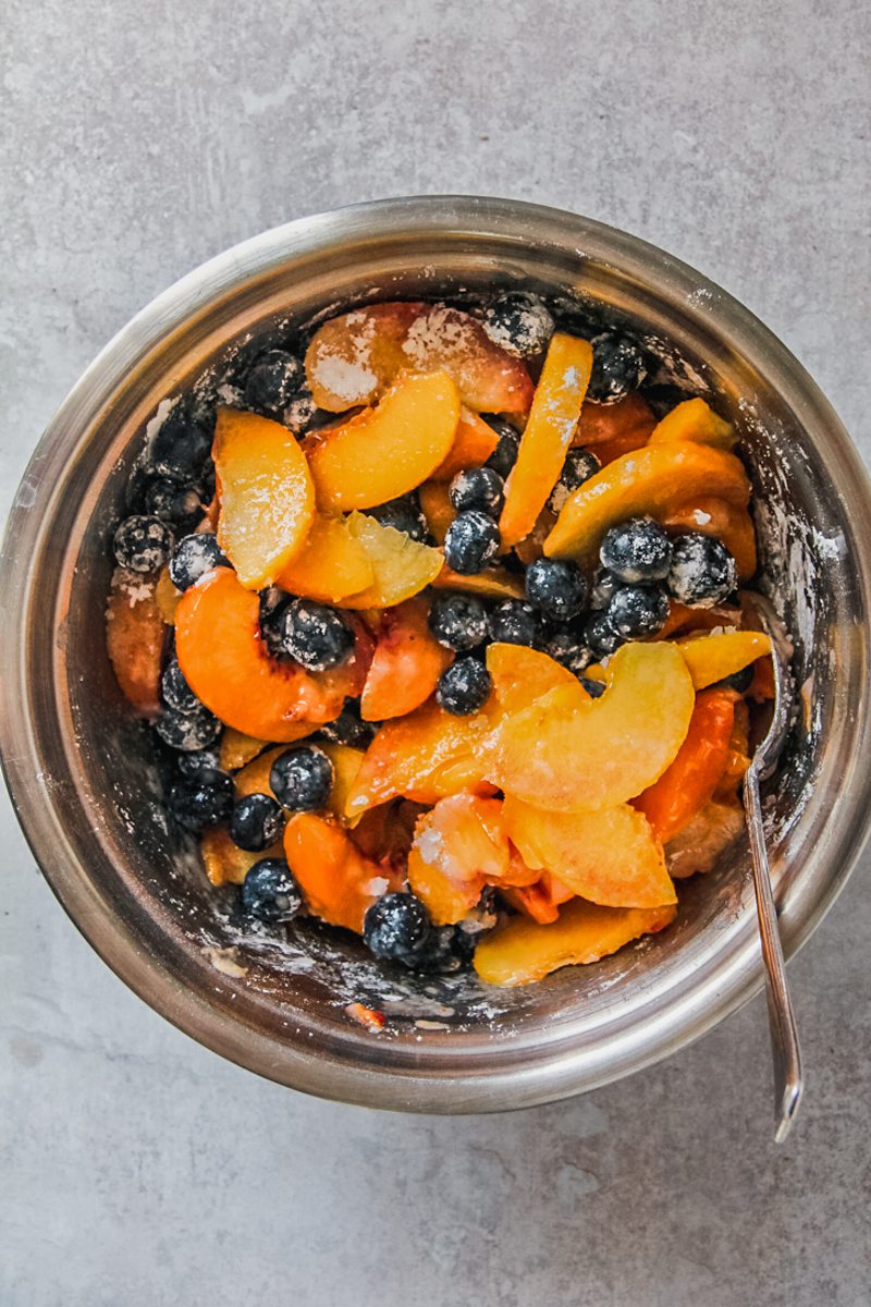 Slices of peaches and blueberries sit stirred with cornflour and sugar in a stainless steel bowl on a gray surface.