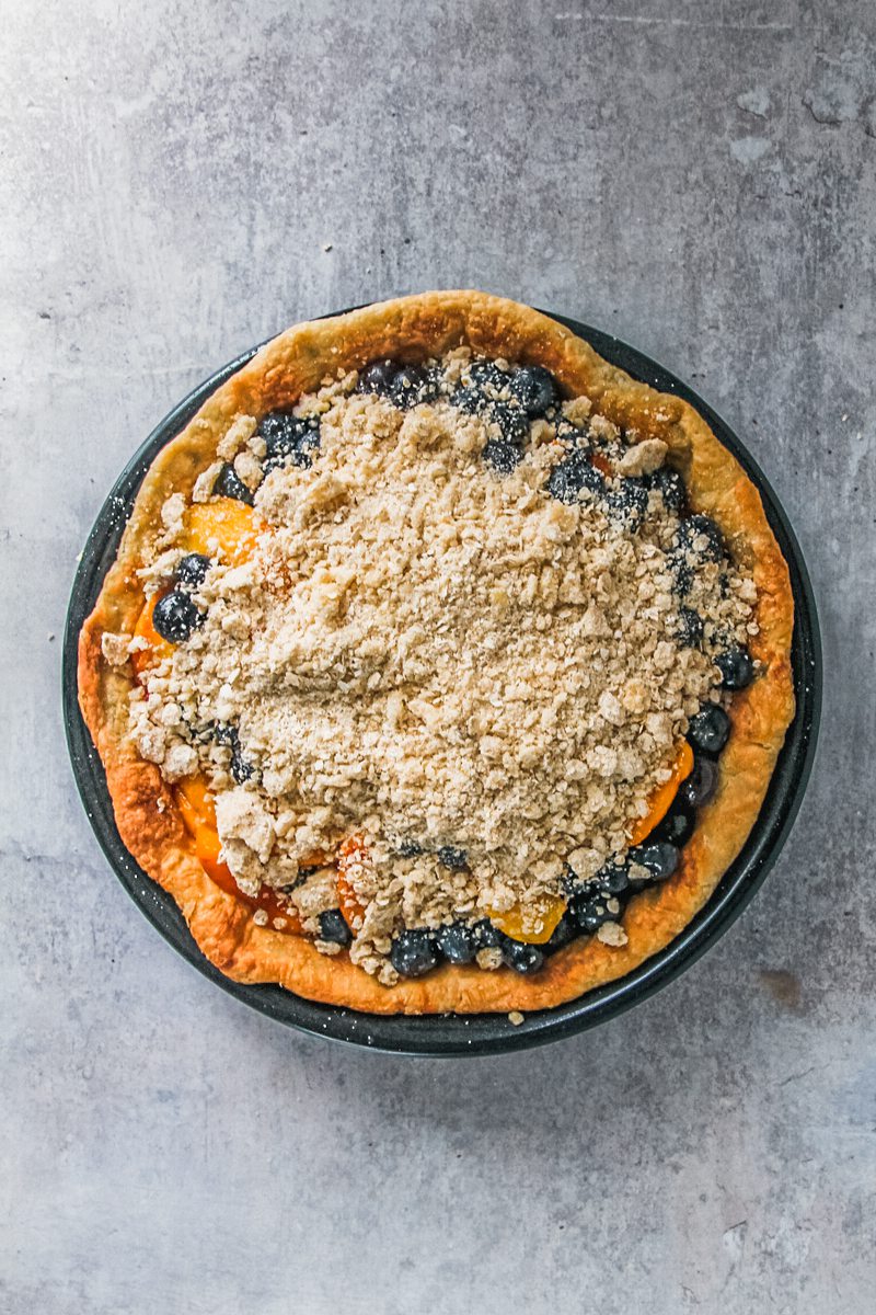 A peach blueberry pie sits par baked in a black metal pie dish with a streusel topping on a gray surface.