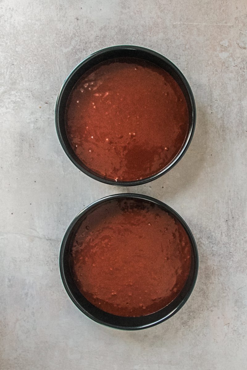 Two metal cake tins with buttermilk chocolate cake batter inside sit on a gray surface.