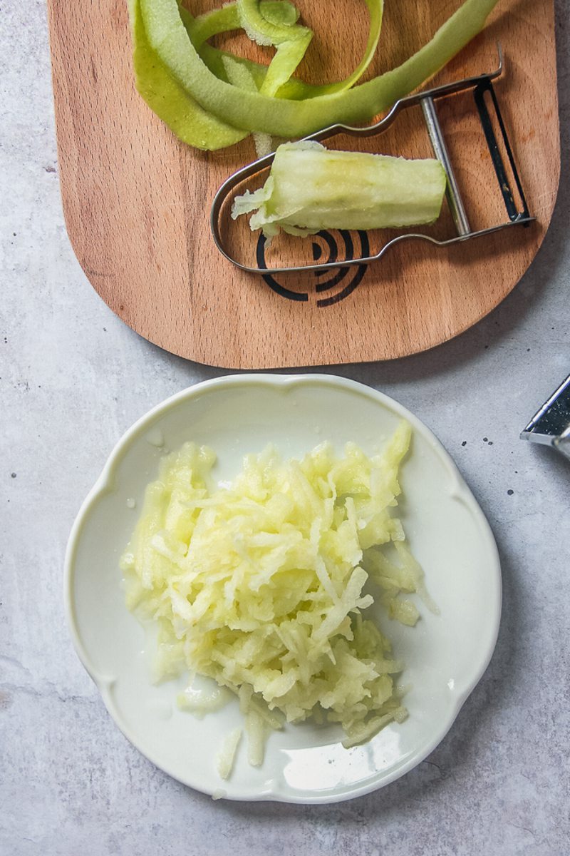 Grated peeled apples sits on top of a white fluted plate on a gray surface with the chopping board, peeler and apple skins beside.