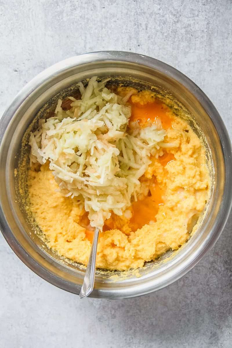 Grated apple sits on top of a cooked cornbread mixture alongside whisked eggs in a stainless steel bowl on a gray surface.