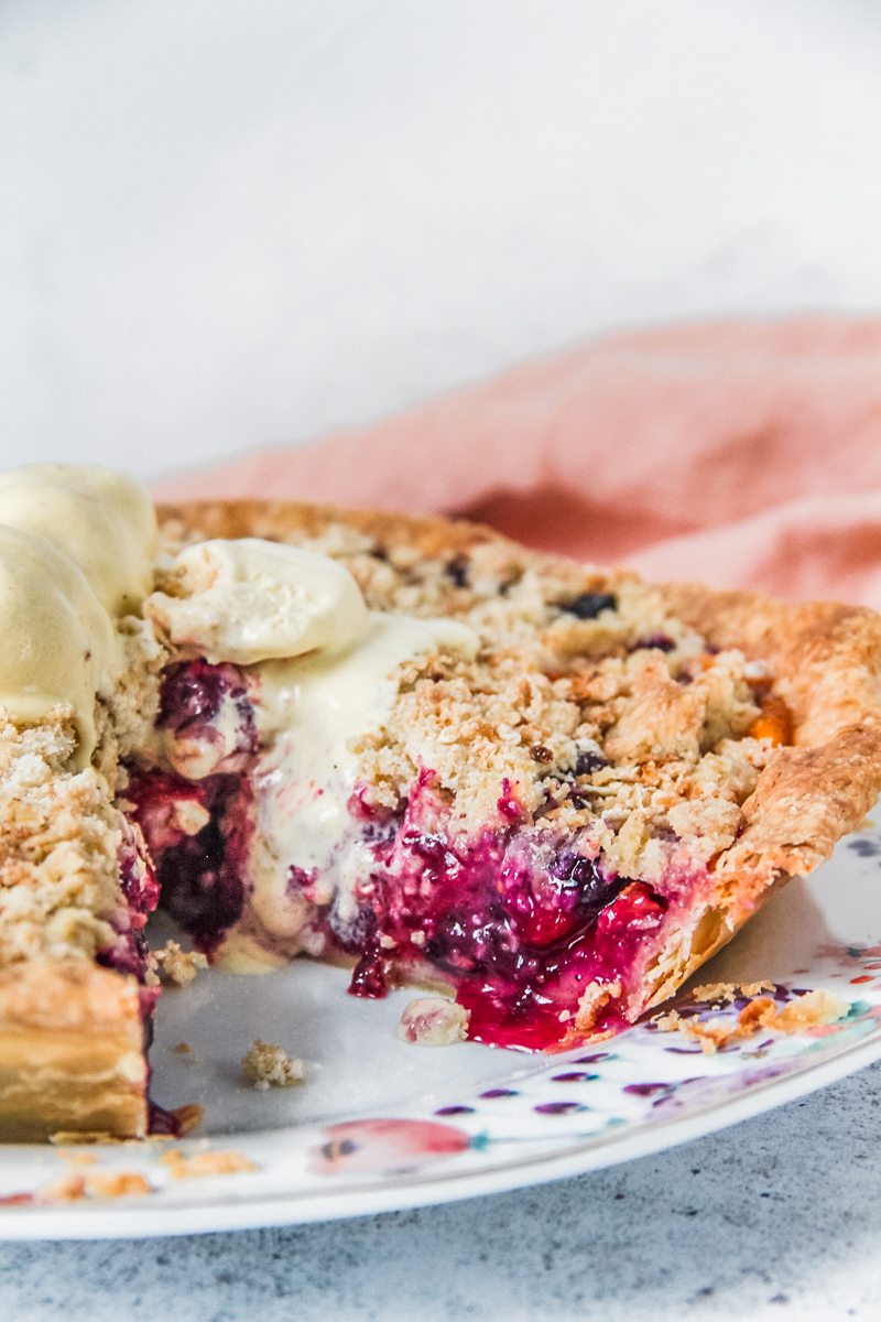 A close up of a peach blueberry streusel pie sits on a plate with melting vanilla ice cream on top and the remaining pie beside on a light gray surface.