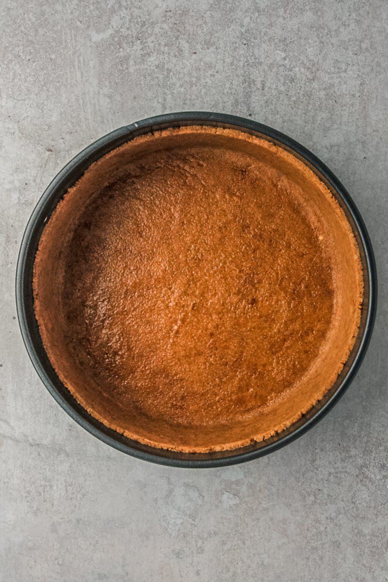 Graham cracker crust sits pressed into a metal cake tin on a gray surface.