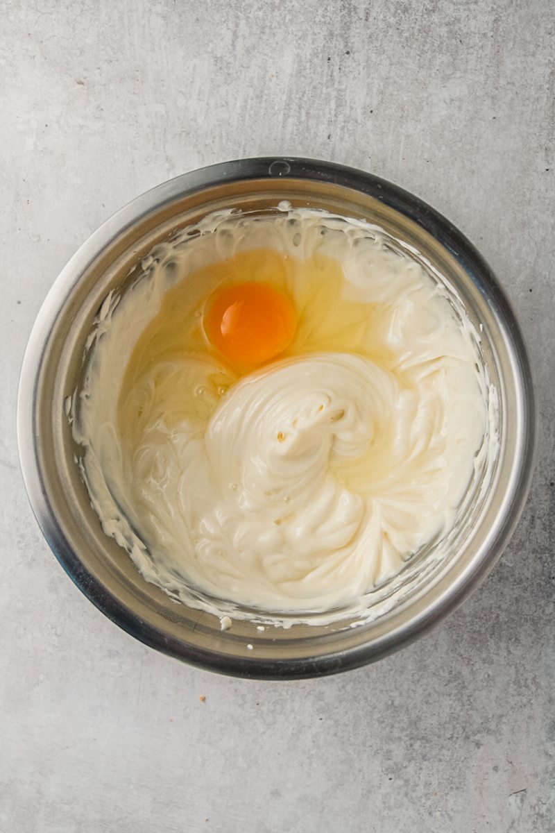 An egg sits on a whipped cheesecake mixture in a stainless steel bowl on a gray surface.