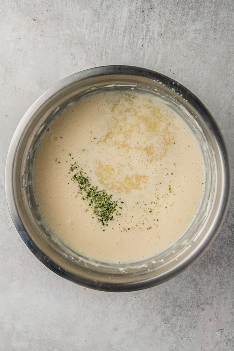 Lime juice and zest sit on top of a whipped cheesecake mixture in a stainless steel bowl on a gray surface.