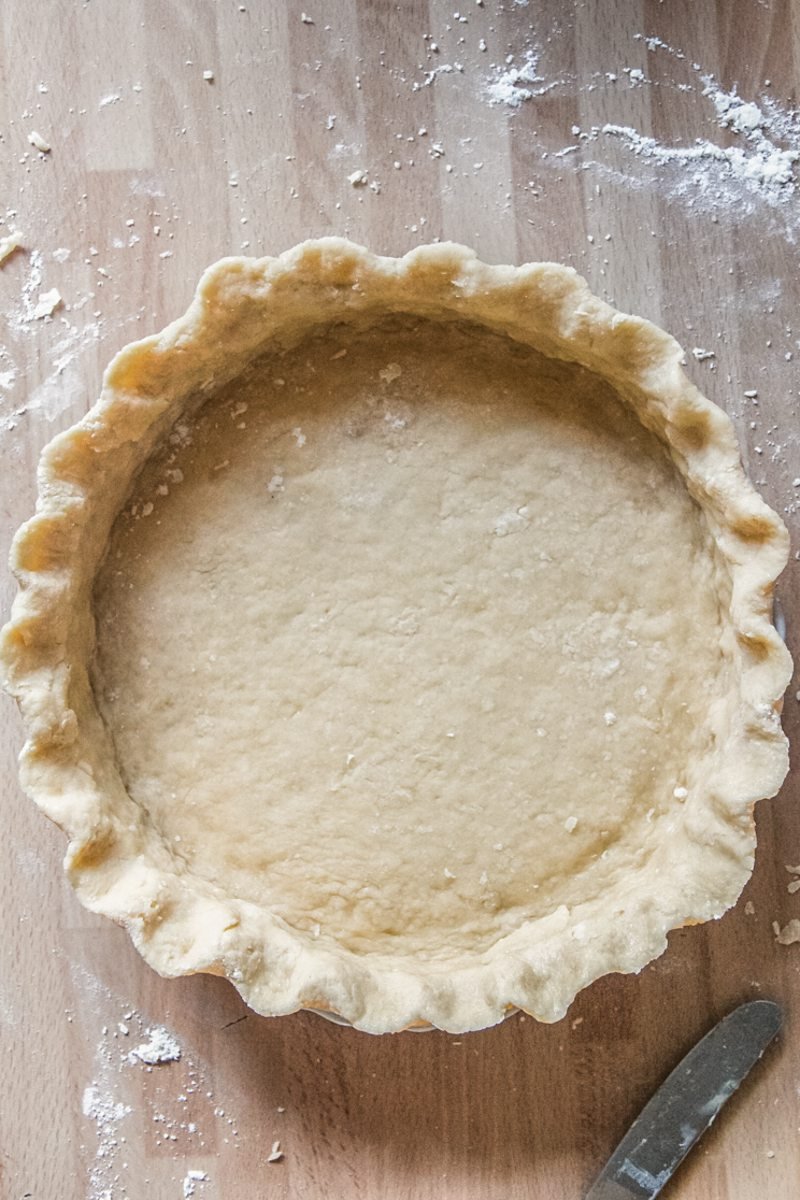 Pie pastry sits pressed and shaped into a pie dish on a wooden benchtop.