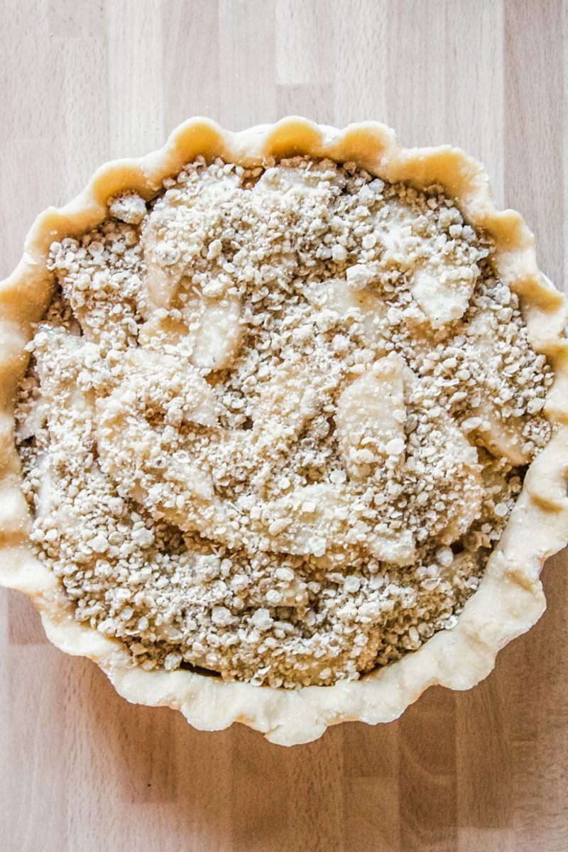 An unbaked pear pie sits on a wooden benchtop with streusel on top.