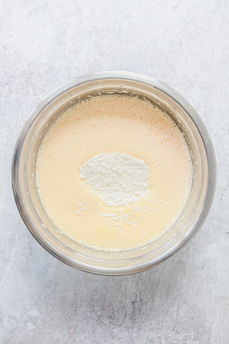 Dry ingredients sink into pale and fluffy whipped eggs in a stainless steel nowl on a gray surface.