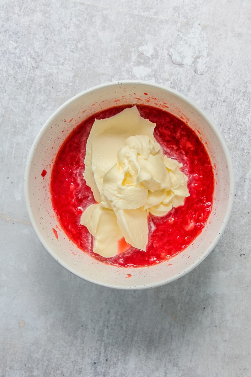 Soft whipped cream sits on top of mashed strawberries in a white ceramic bowl on a gray surface.