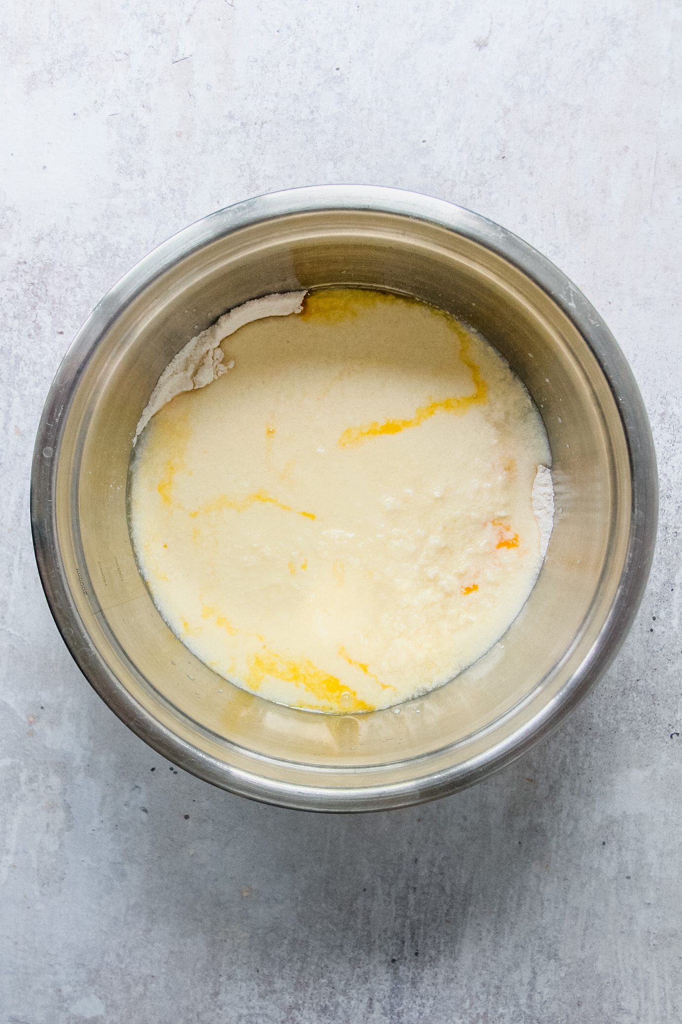 Cream and egg yolks sit on top of dry ingredients in a stainless steel bowl on a gray surface.