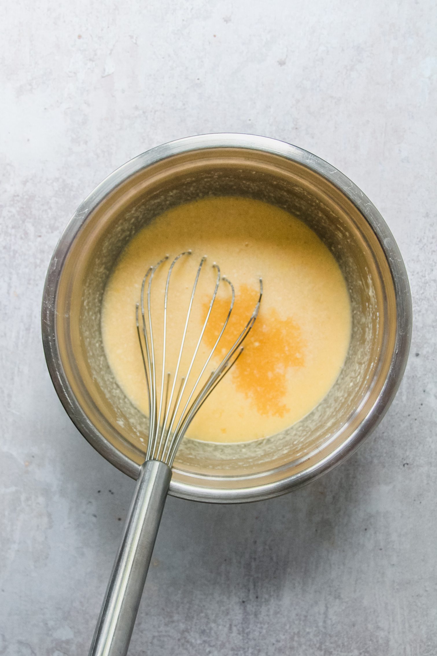 Apple juice sits in a whipped batter mixture in a stainless steel bowl on a gray surface.