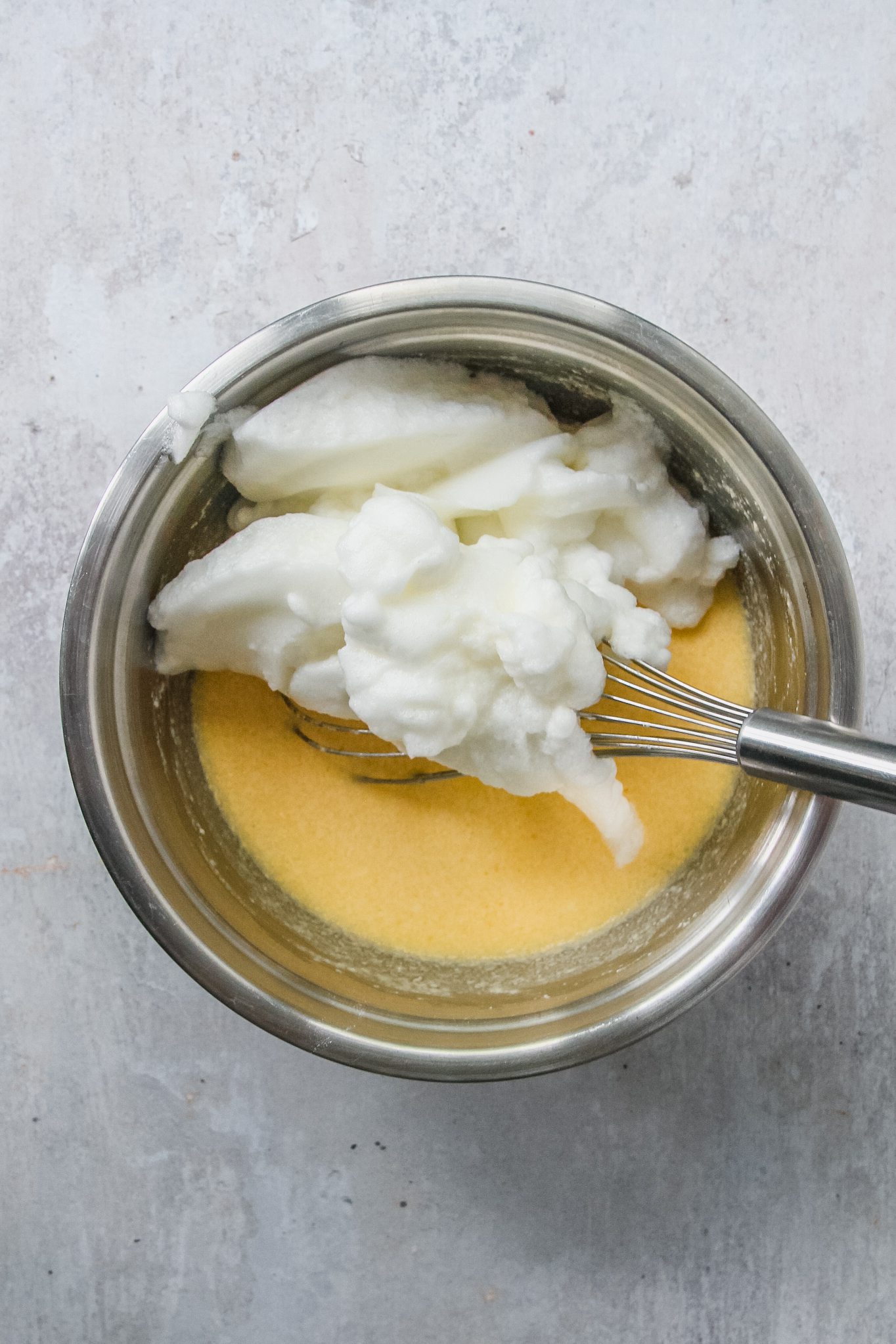 Whipped egg whites sit on top of a batter mixture in a stainless steel bowl on a gray surface.