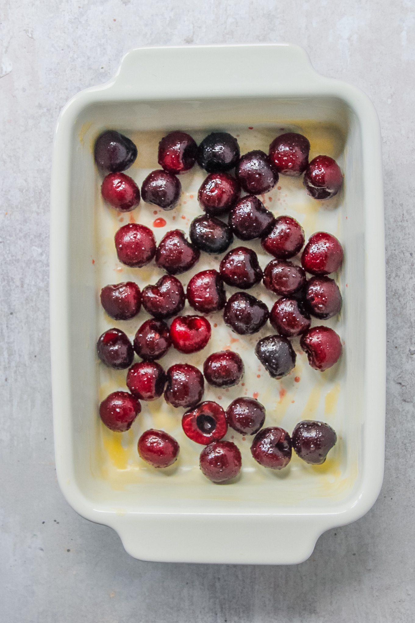 Pitted cherries sit in a buttered rectangle white baking dish on a gray surface.
