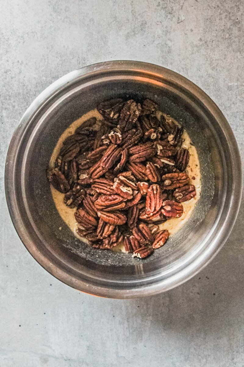 Pecans sit on top of a buttery sugar mixture in a stainless steel bowl on a gray surface.