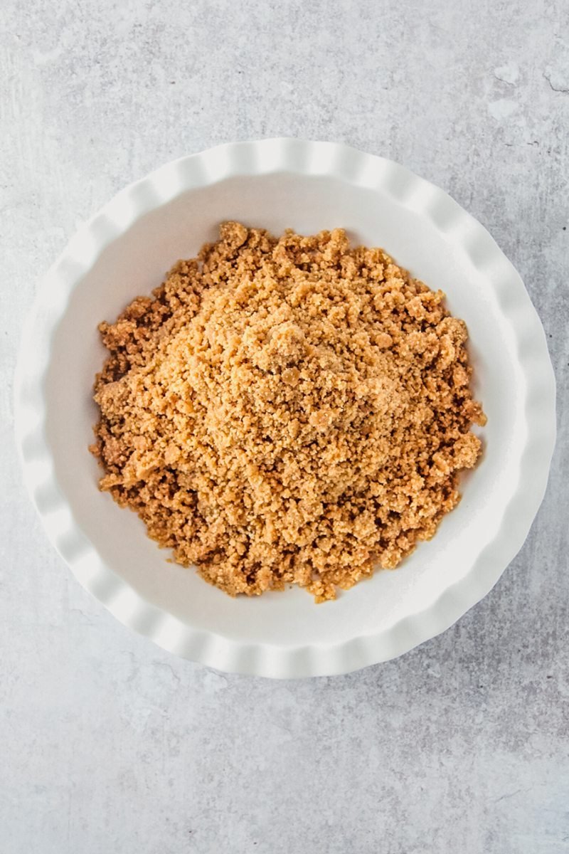 A crushed biscuit base sits in a white ceramic pie dish on a gray surface.